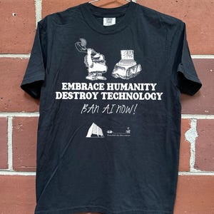 May include: Black t-shirt with white graphic text and illustrations. The text reads "EMBRACE HUMANITY DESTROY TECHNOLOGY BAN AI NOW!" with accompanying images of a cartoon character and a computer.