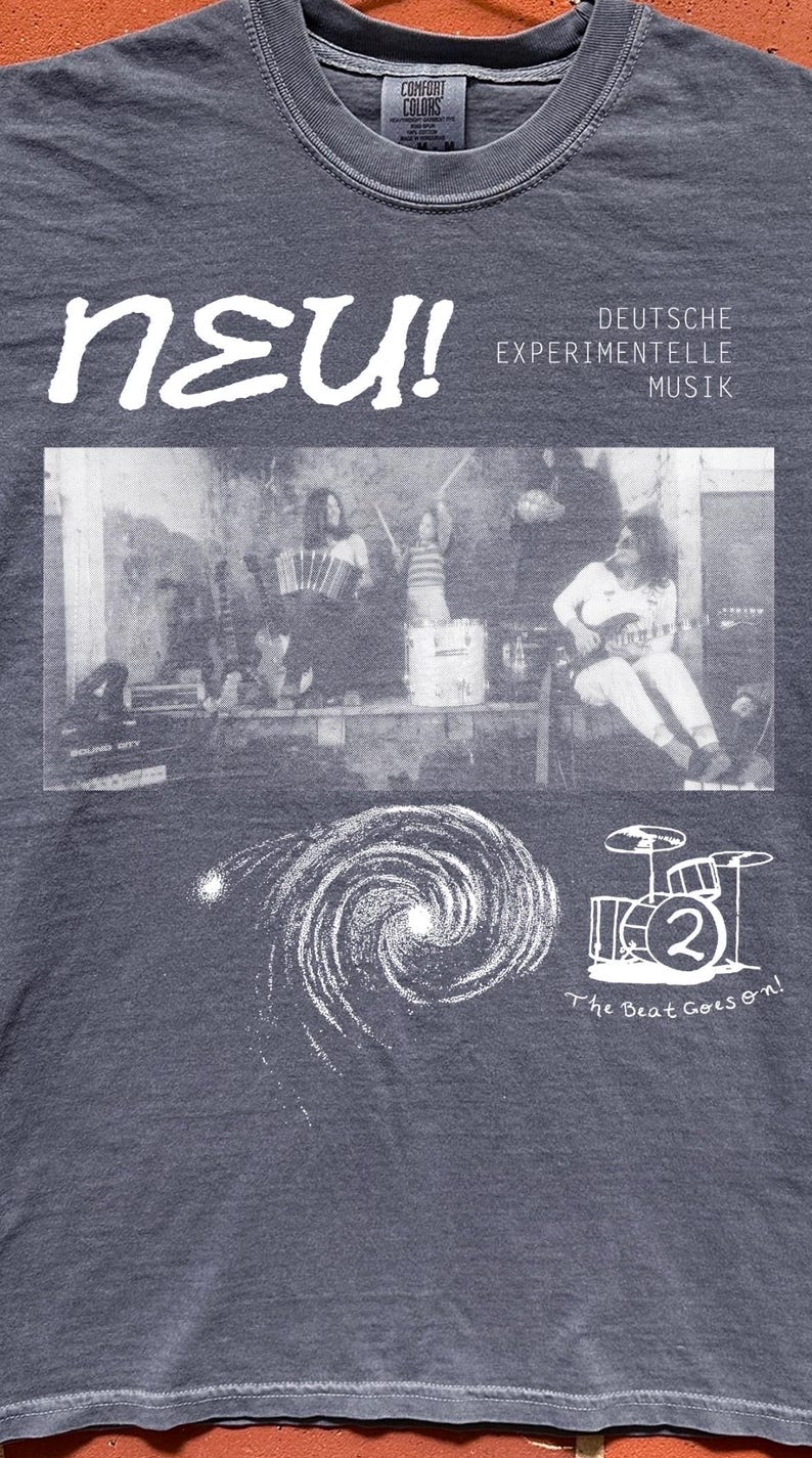 May include: A charcoal gray t-shirt featuring a white graphic print. The design includes the text "neu! DEUTSCHE EXPERIMENTELLE MUSIK," a black and white photograph of a band, a spiral galaxy illustration, a drum kit icon with a "2," and "The Beat Goes on!"