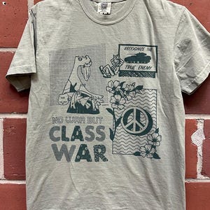 May include: Sage green t-shirt with a graphic design. The design includes the text "NO WAR BUT CLASS WAR", a peace symbol, flowers, and illustrations of a tank and a figure. The shirt is hanging on a metal hanger.