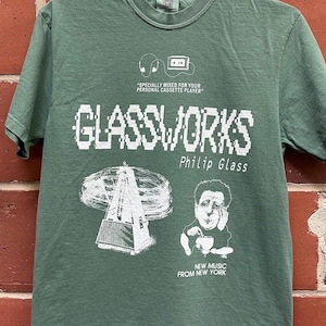 May include: Sage green t-shirt featuring white graphic text and illustrations. The text reads "GLASSWORKS Philip Glass" with additional text and illustrations of a metronome, a cassette player, and a portrait. The shirt is hanging on a metal hanger.