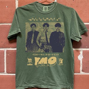 May include: Olive green t-shirt featuring a graphic of three men and the text "MUSIC TO COMPUTE BY". Below the image are Japanese characters and the letters "YMO" in a stylized font. The shirt has a crew neck.