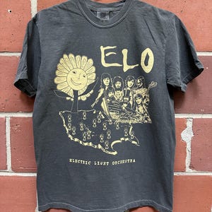 May include: Dark gray t-shirt featuring a gold graphic of a smiling sunflower, band members, and the text "ELO" and "Electric Light Orchestra." The shirt is hanging on a metal hanger against a brick wall.