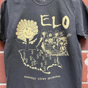 May include: Dark grey t-shirt with a gold graphic of a smiling sunflower, the band ELO, and a map with lightbulb icons. The text "ELECTRIC LIGHT ORCHESTRA" is printed below the map. The shirt is made of a soft material.
