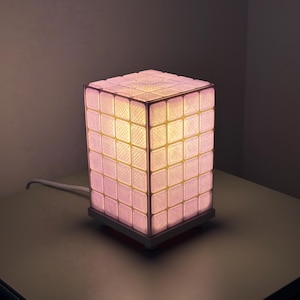 Op de afbeelding: Een kubusvormige lamp met een rasterpatroon, verlicht met een zachte, warme gloed. De buitenkant van de lamp bestaat uit talrijke kleine, vierkante panelen, waardoor een gestructureerd oppervlak ontstaat. De basis is een eenvoudig, rechthoekig ontwerp en een wit snoer loopt van de lamp.