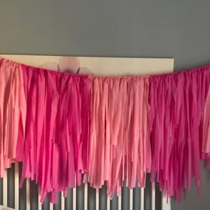 Könnte beinhalten: Eine Girlande aus rosa und fuchsiafarbenem Seidenpapier mit einem mehrschichtigen Fransenmuster. Die Girlande hängt über einem weißen Kinderbett.