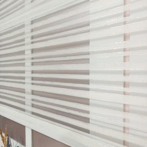 May include: Close-up of a white and grey striped window blind. The blind is made of a sheer, textured material, with alternating opaque and translucent horizontal stripes. The design allows for filtered light and privacy.