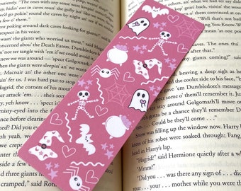 May include: A pink bookmark with a Halloween theme. It features a skeleton, ghosts, bats, spiders, and a cauldron. The bookmark is decorated with white stars and hearts.
