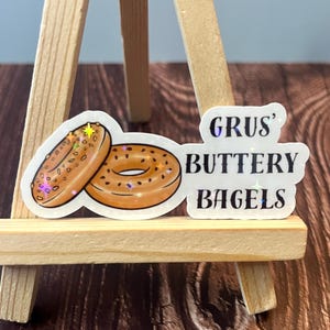 May include: Two cartoon illustrations of bagels with sesame seeds on a white background. The text "GRUS' BUTTERY BAGELS" is written in black with a glittery effect.