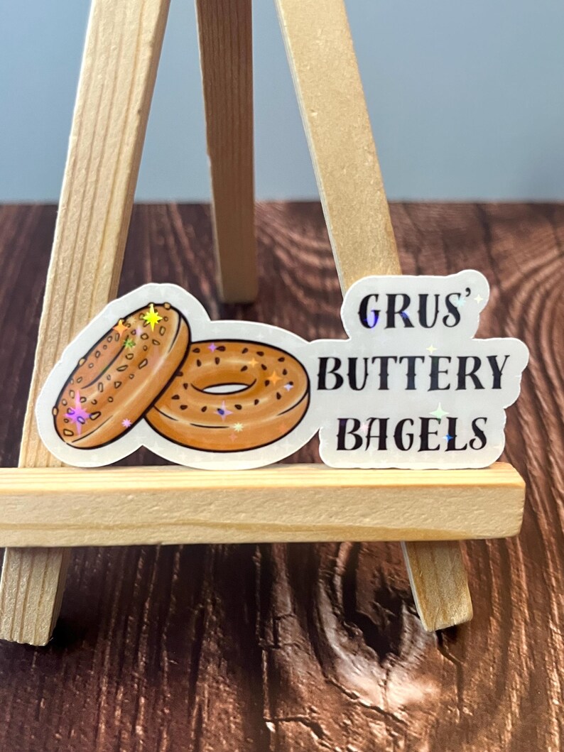 May include: Two cartoon illustrations of bagels with sesame seeds on a white background. The text "GRUS' BUTTERY BAGELS" is written in black with a glittery effect.