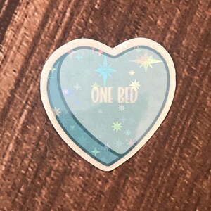 May include: A blue heart-shaped sticker with a holographic finish. The sticker has a white outline and the text "ONE BED" in white lettering.