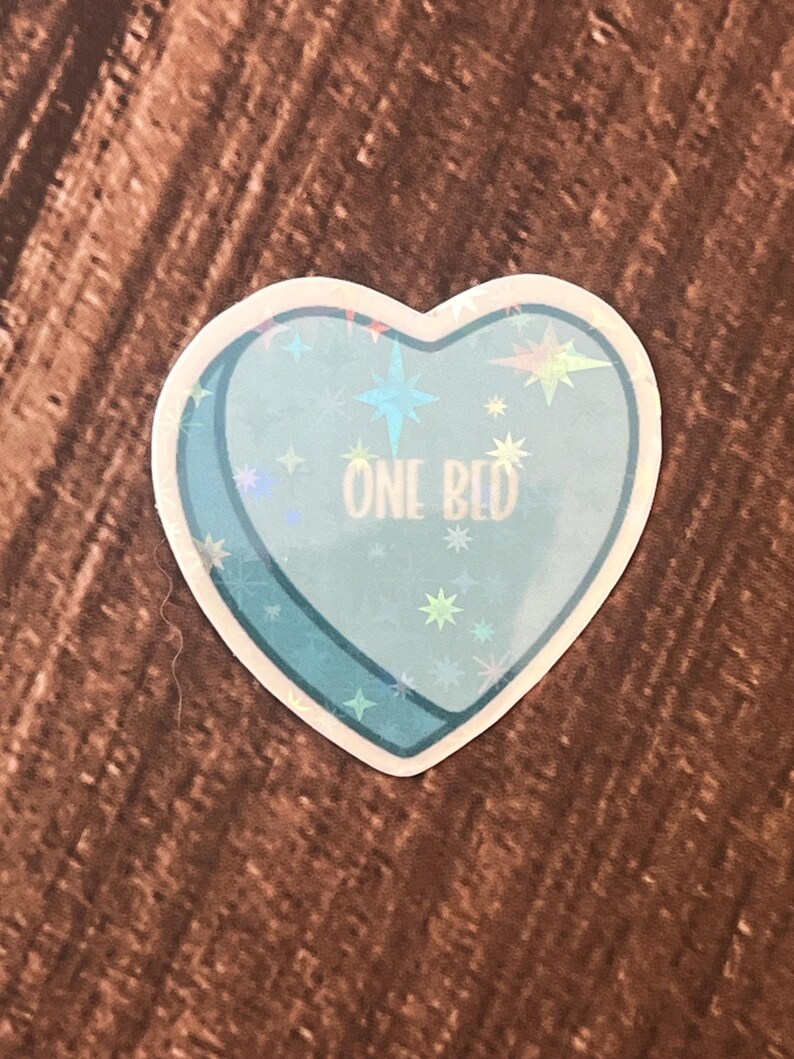 May include: A blue heart-shaped sticker with a holographic finish. The sticker has a white outline and the text "ONE BED" in white lettering.