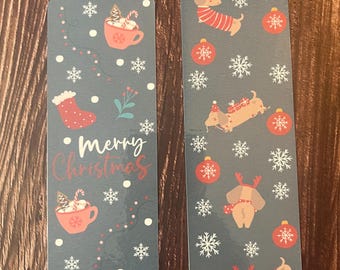 May include: Two Christmas themed bookmarks. The bookmark on the left has a red stocking, a cup of hot chocolate with marshmallows, and the text "Merry Christmas". The bookmark on the right has a dachshund wearing a Santa hat and a red and white striped sweater, a dachshund wearing a red and white striped sweater with reindeer antlers, and a dachshund wearing a red and white striped sweater with a present.