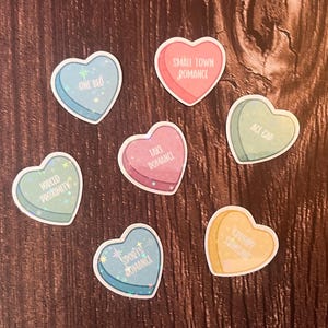 May include: Seven heart-shaped stickers with different sayings. The stickers are in various colors, including pink, blue, green, and yellow. The sayings on the stickers include "One Bed", "Forced Proximity", "Small Town Romance", "Lake Romance", "Act Cap", "Empty Promises", and "Spooky Romance".