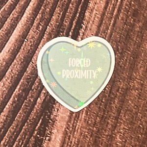 May include: A holographic heart-shaped sticker with a glittery blue background and the text "FORCED PROXIMITY" in white.