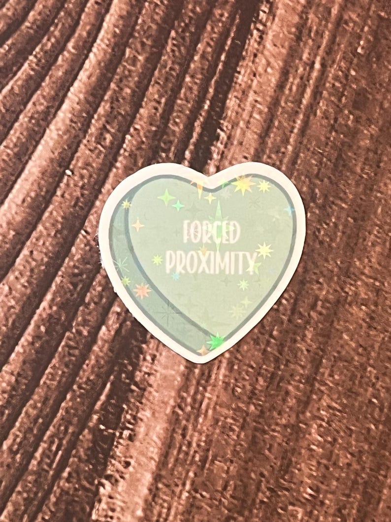 May include: A holographic heart-shaped sticker with a glittery blue background and the text "FORCED PROXIMITY" in white.
