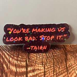 Puede incluir: Una pegatina hologr&aacute;fica negra y roja con el texto "You're making us look bad. Stop it." y la firma "Tairn" en texto m&aacute;s peque&ntilde;o.