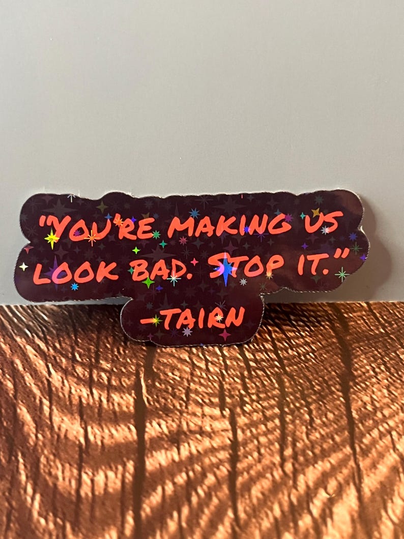 Puede incluir: Una pegatina hologr&aacute;fica negra y roja con el texto "You're making us look bad. Stop it." y la firma "Tairn" en texto m&aacute;s peque&ntilde;o.