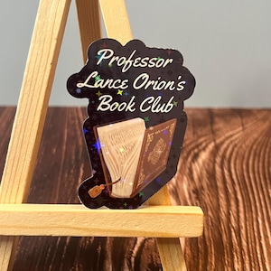May include: A black and gold holographic sticker with the text "Professor Lance Orion's Book Club" and an image of an open book with a bookmark.