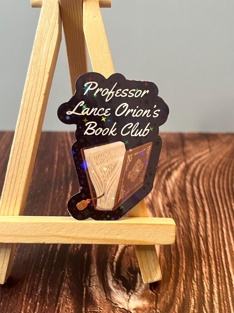 May include: A black and gold holographic sticker with the text "Professor Lance Orion's Book Club" and an image of an open book with a bookmark.