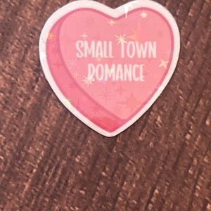 May include: A pink heart-shaped sticker with the text "SMALL TOWN ROMANCE" in white lettering. The sticker has a glittery finish and is decorated with small white stars.