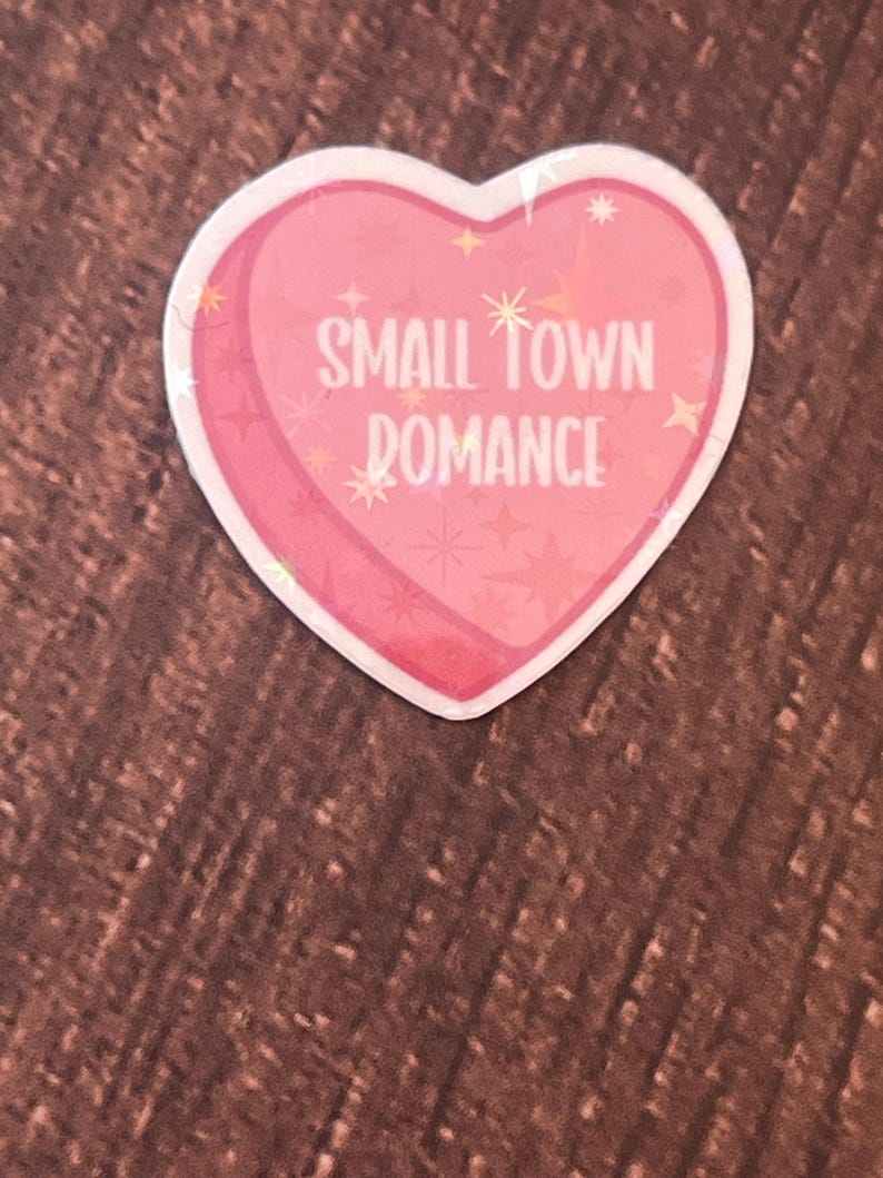 May include: A pink heart-shaped sticker with the text "SMALL TOWN ROMANCE" in white lettering. The sticker has a glittery finish and is decorated with small white stars.