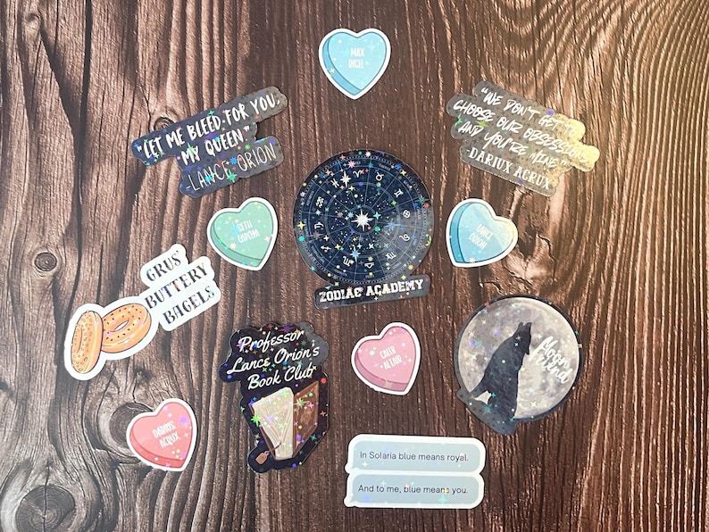May include: A collection of stickers featuring various designs, including a zodiac wheel, a wolf howling at the moon, a book club logo, and heart-shaped stickers with text. The stickers are on a wooden surface.