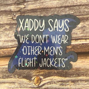 May include: A black and blue sticker with a glittery finish. The sticker features a quote that reads "Xaddy Says "We Don't Wear Other Men's Flight Jackets".