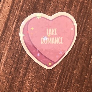 May include: Pink heart-shaped sticker with a glittery finish. The sticker says "FAKE ROMANCE" in white text.