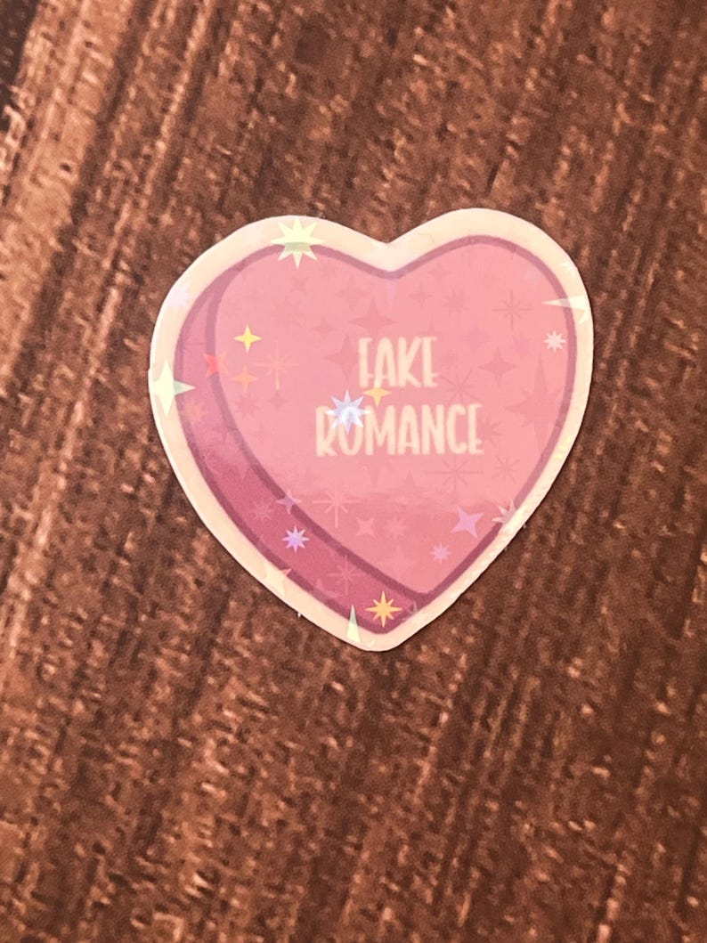 May include: Pink heart-shaped sticker with a glittery finish. The sticker says "FAKE ROMANCE" in white text.