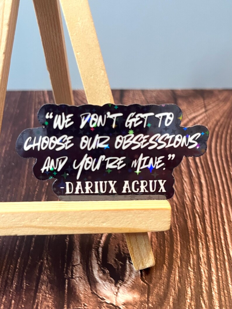 May include: A black and white sticker with a quote from the book "A Court of Thorns and Roses" by Sarah J. Maas. The quote reads: "We don't get to choose our obsessions and you're mine." The quote is attributed to "DARIUX ACRUX".