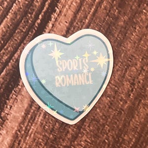 May include: A blue heart-shaped sticker with a holographic finish. The sticker has the text "SPORTS ROMANCE" in white letters.