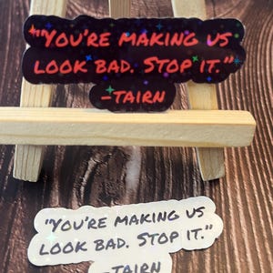 Puede incluir: Dos pegatinas con fondo negro y purpurina iridiscente. El texto en las pegatinas dice "You're making us look bad. Stop it." y est&aacute; escrito en blanco, con una fuente de letra manuscrita. El texto "TAIRN" est&aacute; escrito en rojo debajo del texto principal.