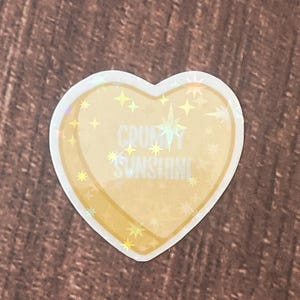 May include: A heart-shaped sticker with a holographic finish. The sticker is yellow with a white outline and features the text "CRUSHY SUNSHINE" with white stars.