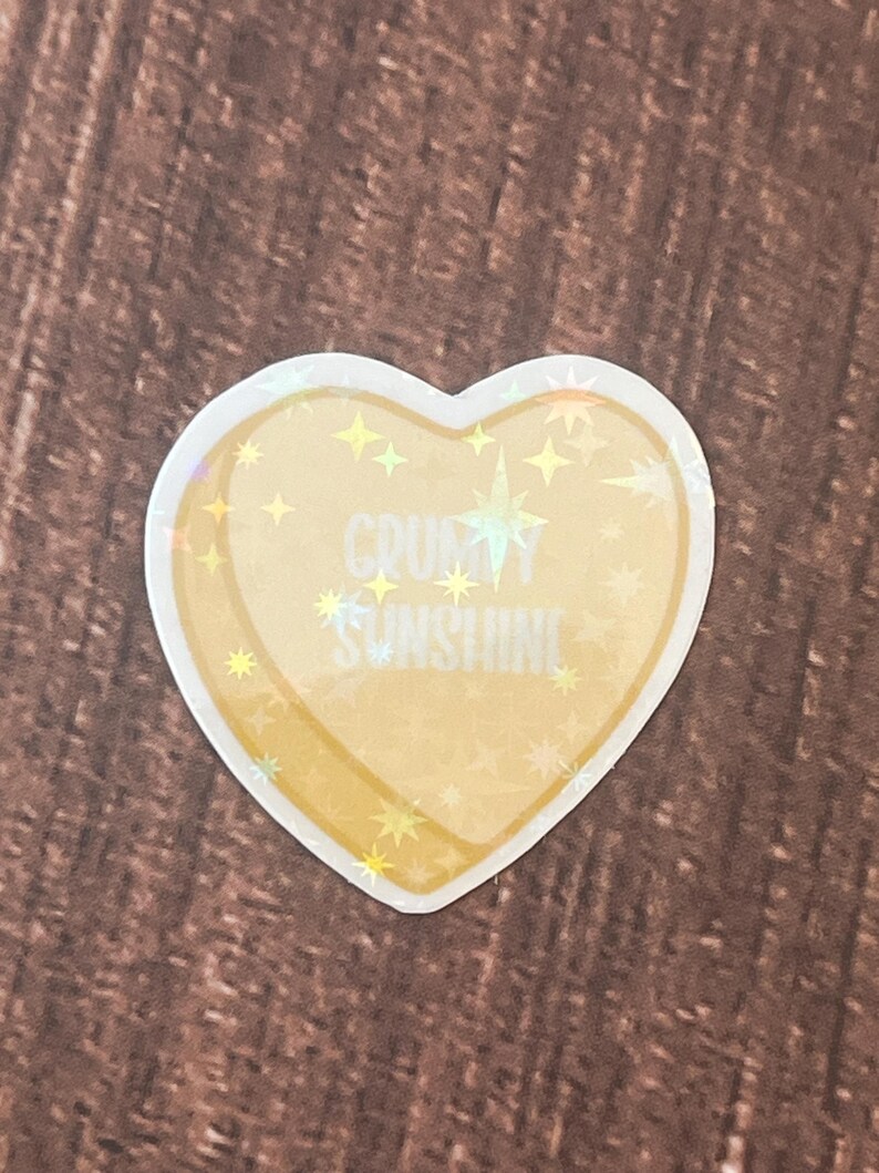 May include: A heart-shaped sticker with a holographic finish. The sticker is yellow with a white outline and features the text "CRUSHY SUNSHINE" with white stars.
