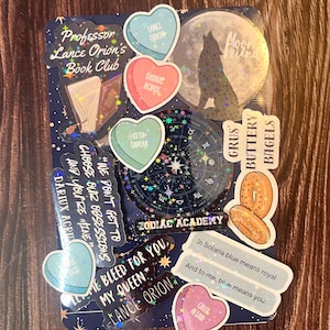 May include: A collection of holographic stickers featuring a wolf howling at the moon, a book, a zodiac wheel, and various heart-shaped stickers with text like "Lance Orion" and "Darius Acrux". The stickers are on a dark blue background with stars.