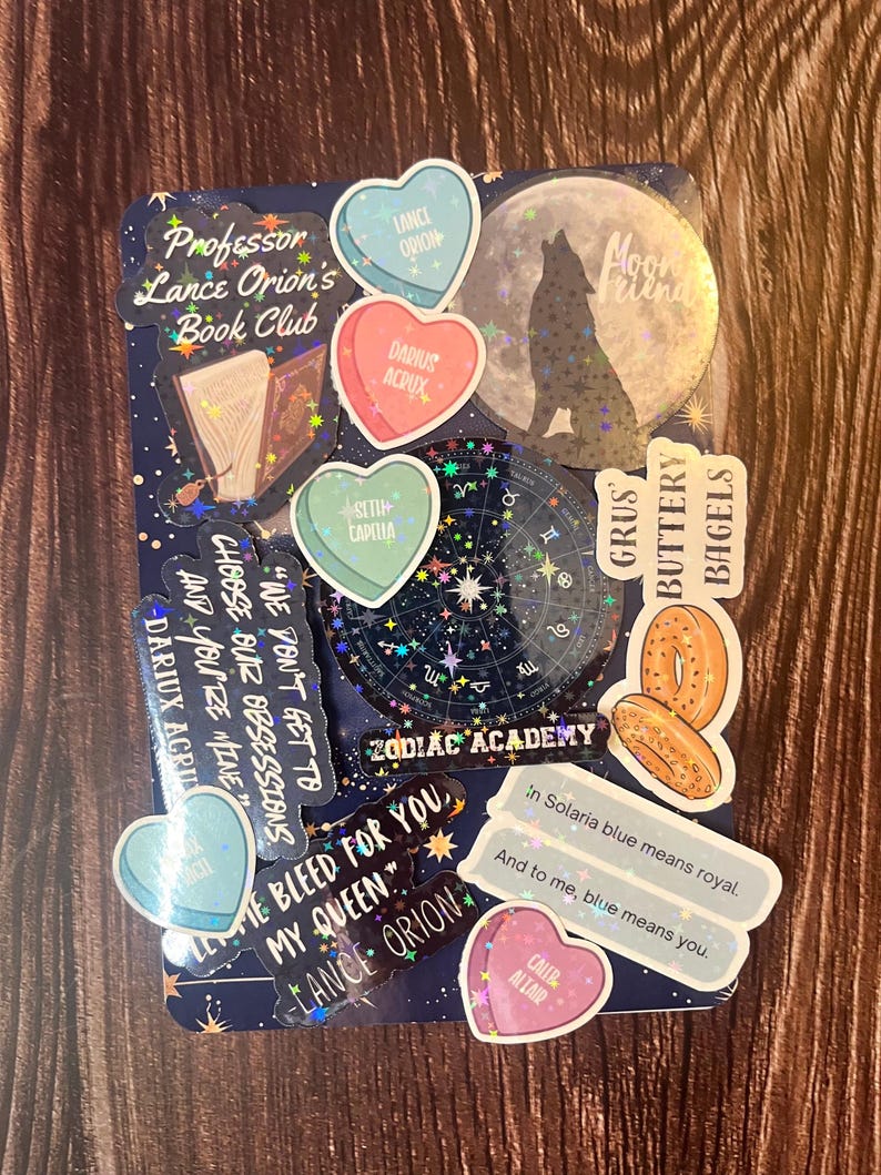 May include: A collection of holographic stickers featuring a wolf howling at the moon, a book, a zodiac wheel, and various heart-shaped stickers with text like "Lance Orion" and "Darius Acrux". The stickers are on a dark blue background with stars.