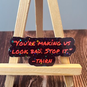 Puede incluir: Una pegatina negra con una cita roja y blanca que dice "You're making us look bad. Stop it." con el nombre "Tairn" escrito debajo de la cita. La pegatina est&aacute; en un peque&ntilde;o caballete de madera.
