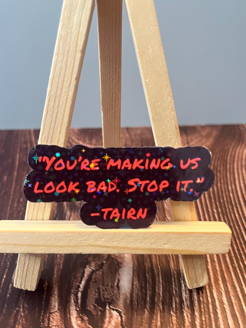 Puede incluir: Una pegatina negra con una cita roja y blanca que dice "You're making us look bad. Stop it." con el nombre "Tairn" escrito debajo de la cita. La pegatina est&aacute; en un peque&ntilde;o caballete de madera.