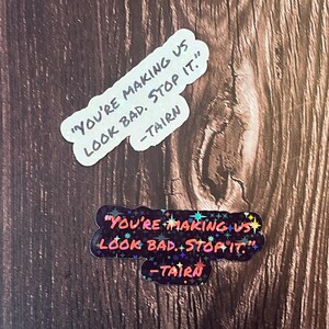 Puede incluir: Dos pegatinas con el texto "You're making us look bad. Stop it." y la firma "Tairn". La pegatina superior es blanca con texto negro. La pegatina inferior es negra con purpurina de colores y texto rojo.