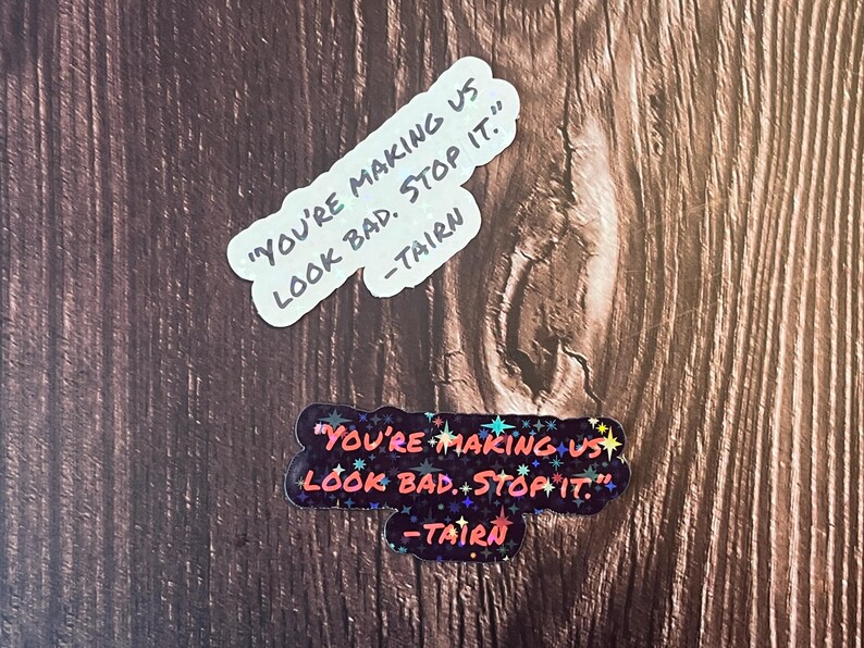 Puede incluir: Dos pegatinas con el texto "You're making us look bad. Stop it." y la firma "Tairn". La pegatina superior es blanca con texto negro. La pegatina inferior es negra con purpurina de colores y texto rojo.