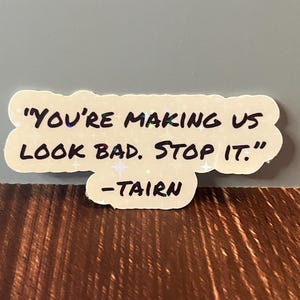 Puede incluir: Una pegatina blanca con una cita manuscrita en negro que dice "You're making us look bad. Stop it." -Tairn