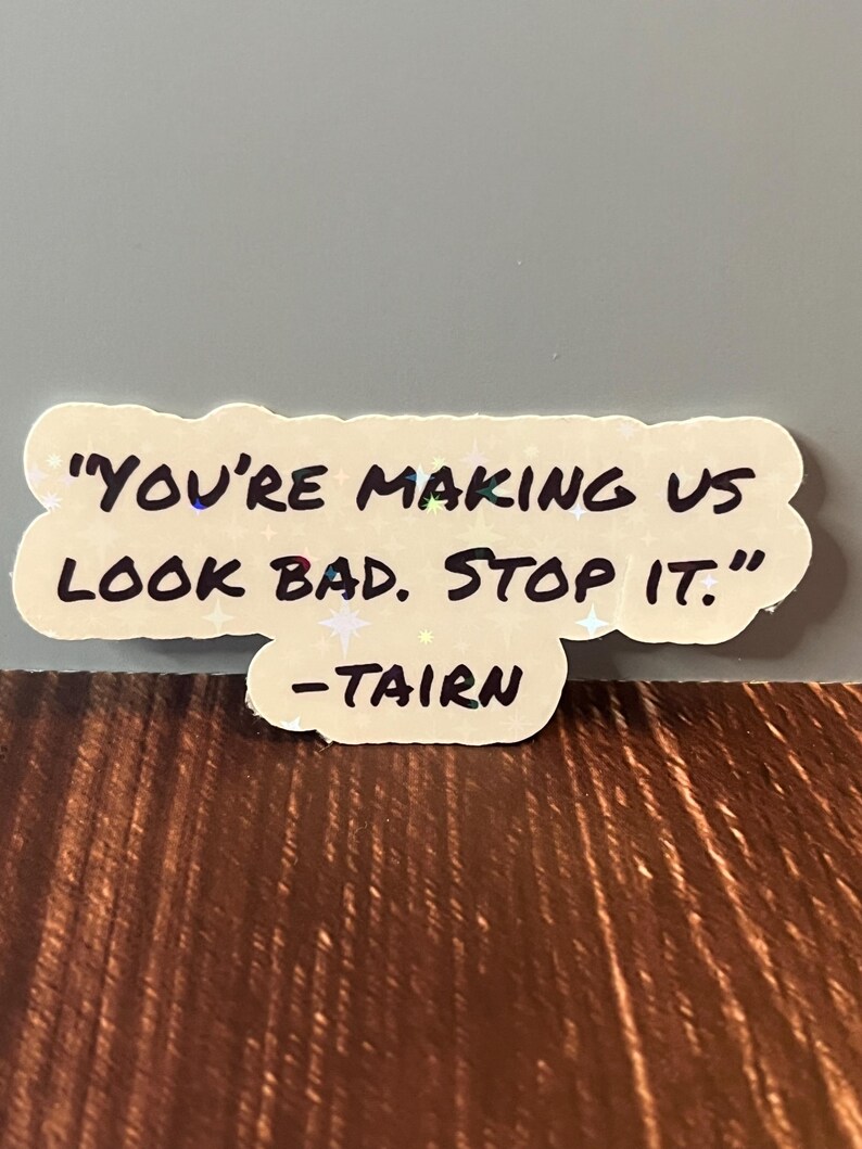 Puede incluir: Una pegatina blanca con una cita manuscrita en negro que dice "You're making us look bad. Stop it." -Tairn