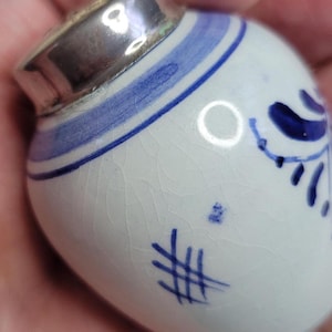 Może przedstawiać: Mały, biały ceramiczny słoik z niebieskimi kwiatowymi wzorami i srebrną metalową pokrywką. Słoik posiada ręcznie malowane niebieskie akcenty i spękaną powierzchnię, prezentując tradycyjną estetykę.