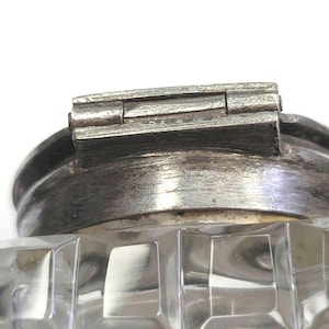 May include: Close-up of a clear, faceted glass jar with a silver-coloured metal lid. The lid has a hinged design and a brushed metal finish. The jar's body features a repeating geometric pattern.