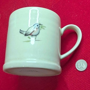 Könnte beinhalten: Cremefarbene Keramiktasse mit einem blau-weißen Vogelmotiv, der einen grünen Zweig hält. Die Tasse hat einen gebogenen Henkel und einen ausgestellten Boden. Eine Münze liegt neben der Tasse zum Größenvergleich. Die Tasse steht auf rotem Untergrund.