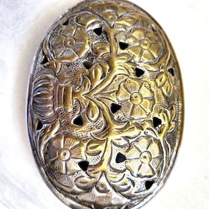 Puede incluir: Una hebilla de cinturón ovalada de latón con un intrincado diseño calado de flores y hojas en relieve. La superficie metálica muestra un acabado dorado pulido con áreas empotradas oscurecidas que resaltan los detalles del patrón floral.