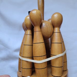 Puede incluir: Un juego de bolos de madera con acabado marr&oacute;n y rayas negras. Los bolos est&aacute;n dispuestos en c&iacute;rculo y unidos con una banda el&aacute;stica. Los bolos est&aacute;n sobre una base de madera.