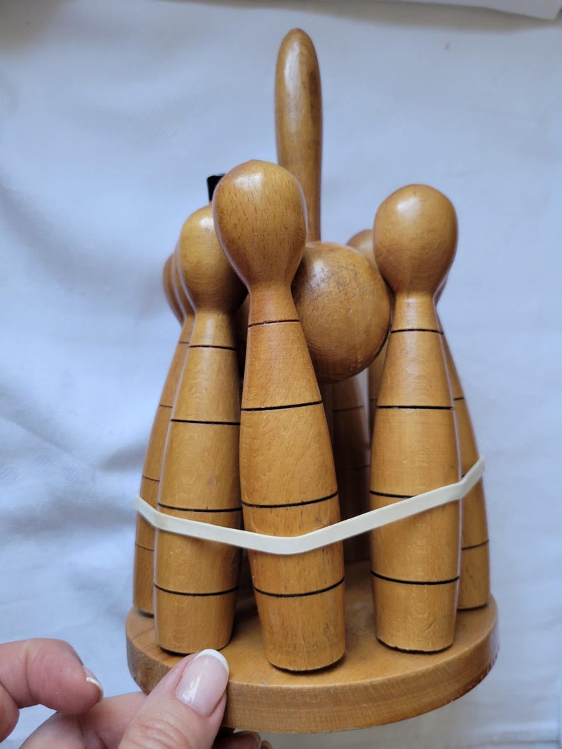 Puede incluir: Un juego de bolos de madera con acabado marr&oacute;n y rayas negras. Los bolos est&aacute;n dispuestos en c&iacute;rculo y unidos con una banda el&aacute;stica. Los bolos est&aacute;n sobre una base de madera.