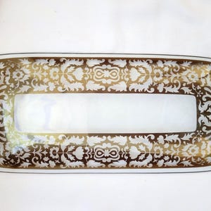 Può includere: Vassoio rettangolare con centro bianco e bordo decorativo. Il bordo presenta un motivo floreale dorato e bianco. Il vassoio ha una finitura lucida ed è probabilmente realizzato in vetro o ceramica.