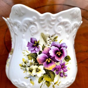 May include: A white porcelain jug featuring a scalloped rim and embossed scrollwork details. The front is decorated with a colourful floral arrangement of purple and yellow pansies, green leaves, and small white blossoms with gold trim.